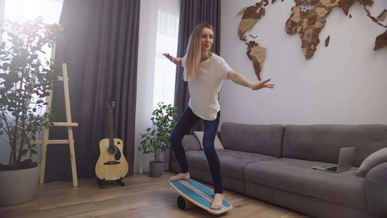 Woman balancing on a balance board indoors with arms spread for stability.