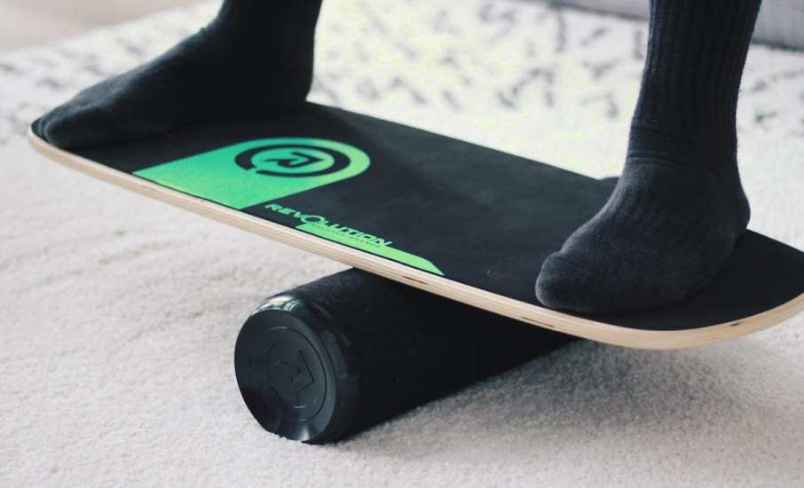 Feet in socks balancing on a black and green Revolution balance board indoors.