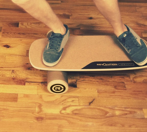 Person training on a Revolution EcoSeries balance board with a roller on a wood floor.